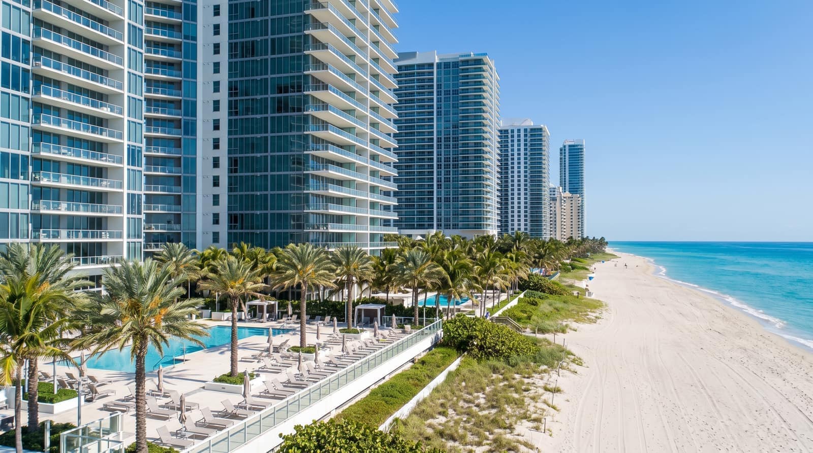 Tall Florida oceanfront condominium towers rising along a wide sandy beach with palms and a pool deck