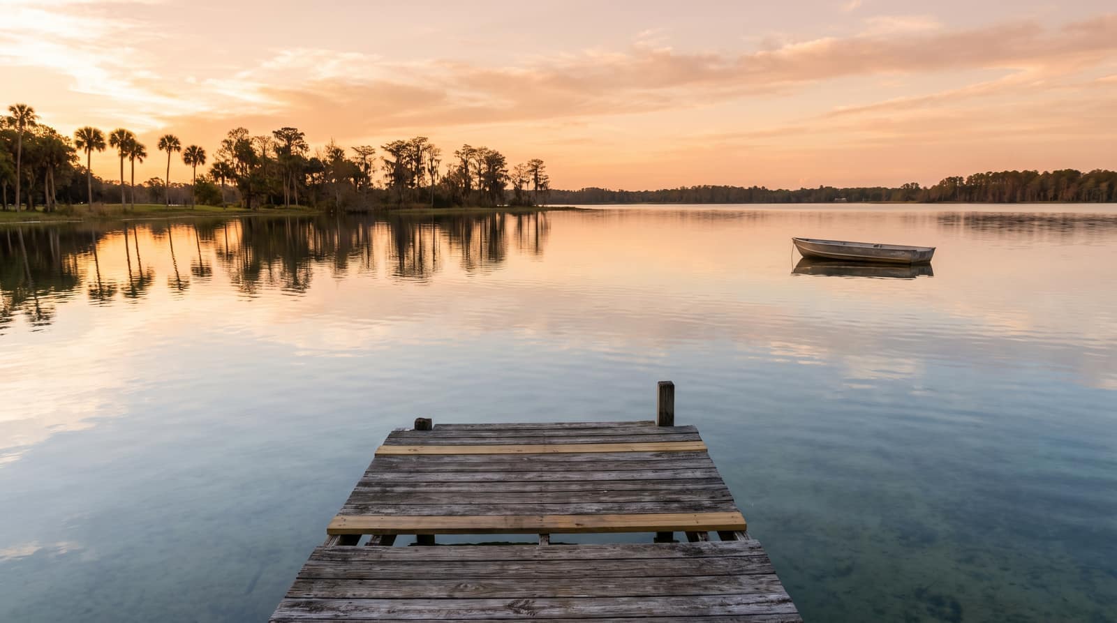A wide calm Florida lake at sunrise with a single fishing boat in the distance and a wooden dock