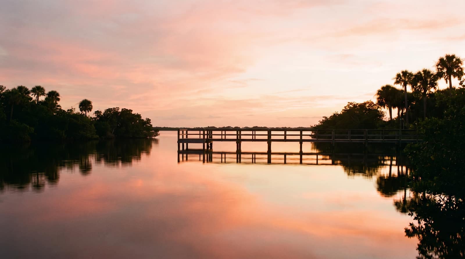 The calm Indian River Lagoon at sunset with a long wooden community pier over still water and silhouetted palms