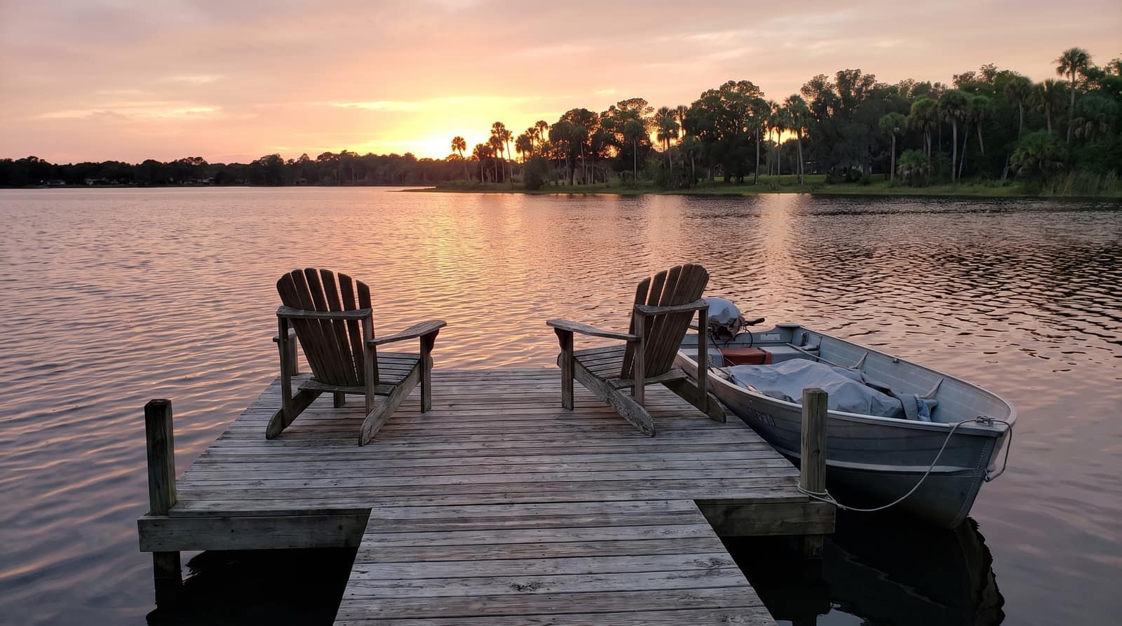 A calm Central Florida lake at sunset seen from a private wooden dock with two Adirondack chairs and a small moored boat