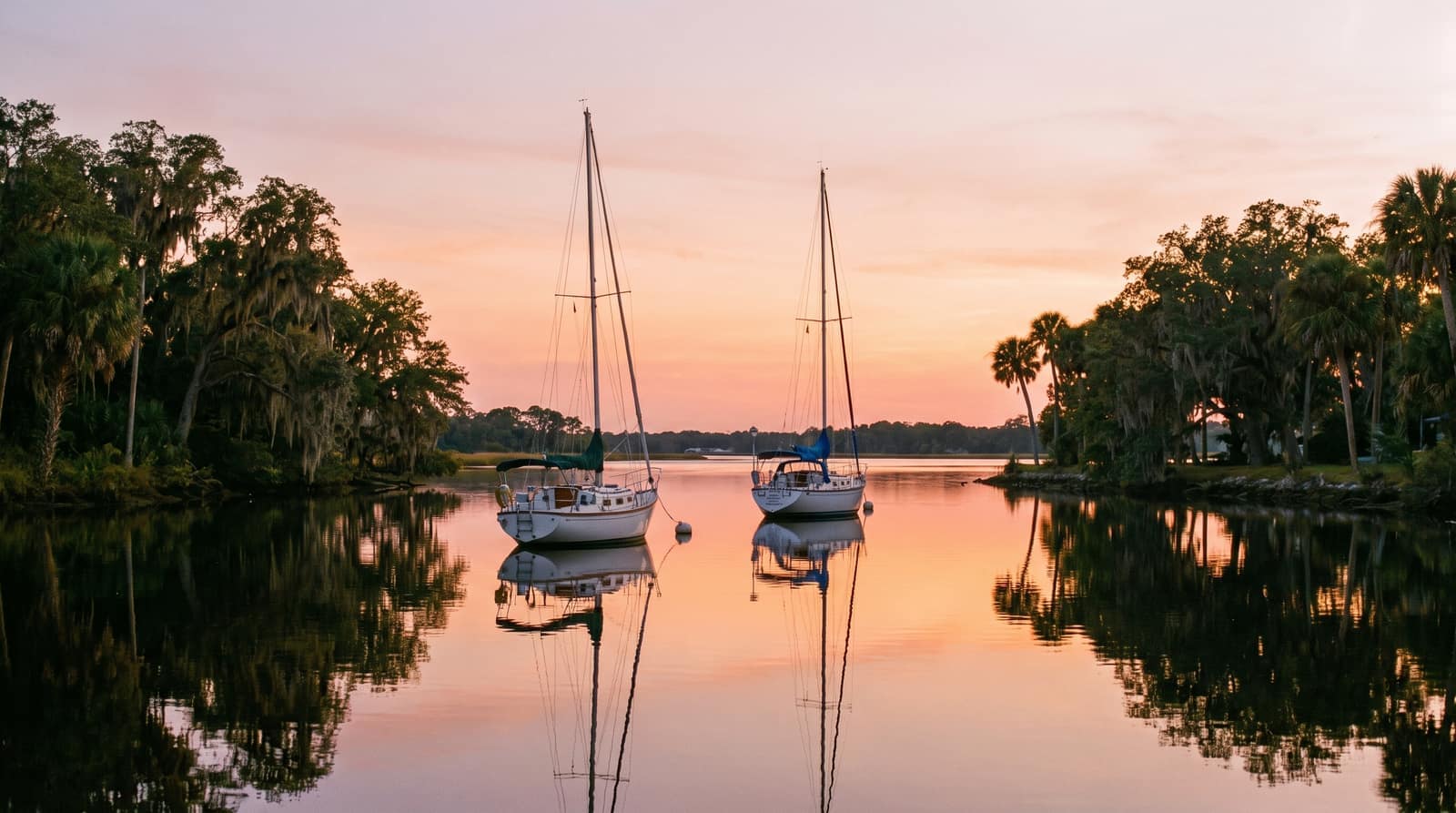 The wide Halifax River at sunset with sailboats anchored on calm water and oak-and-palm-lined banks